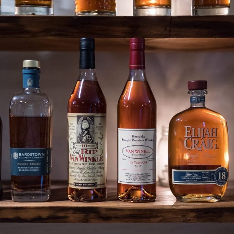 Four bottles of premium bourbon whiskey, including Van Winkle and Elijah Craig, displayed on a wooden shelf.