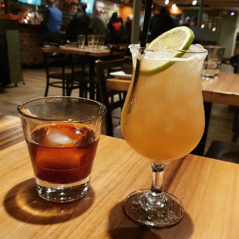 Two drinks, a dark cocktail and a margarita, on a wooden table at Fox's Den gastropub.
